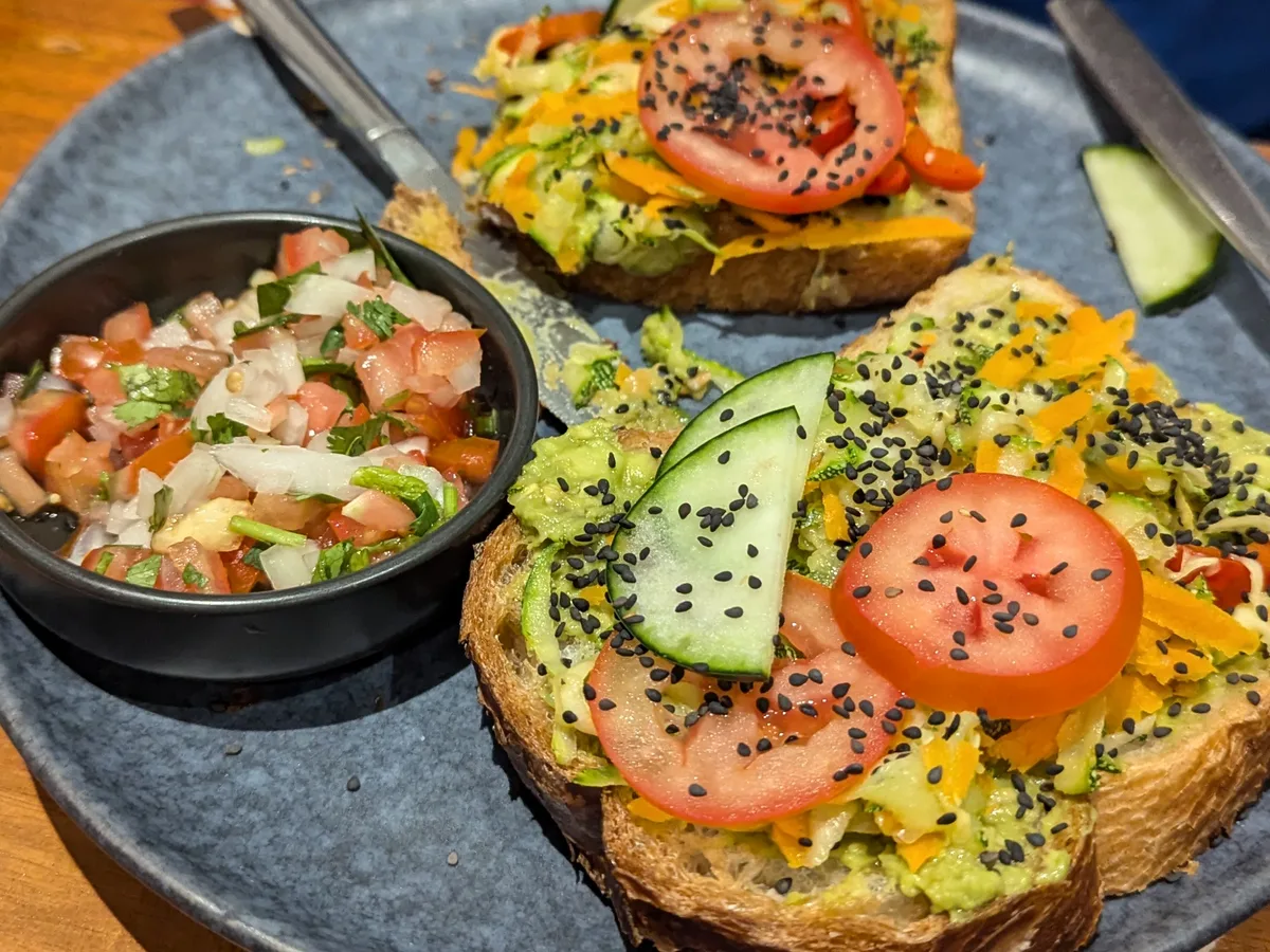 Zwei Scheiben Avocado-Toast mit Tomate, Gurke und schwarzem Sesam auf einem blauen Teller