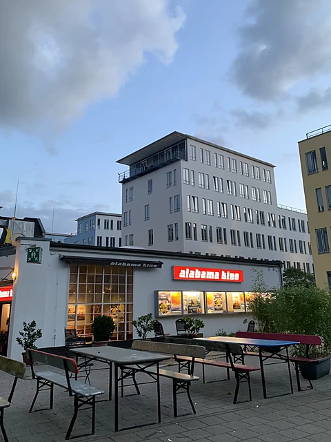 Kinogebäude in der Dämmerung mit rot leuchtendem Schriftzug, davor Biertischgarnituren auf gepflastertem Hof