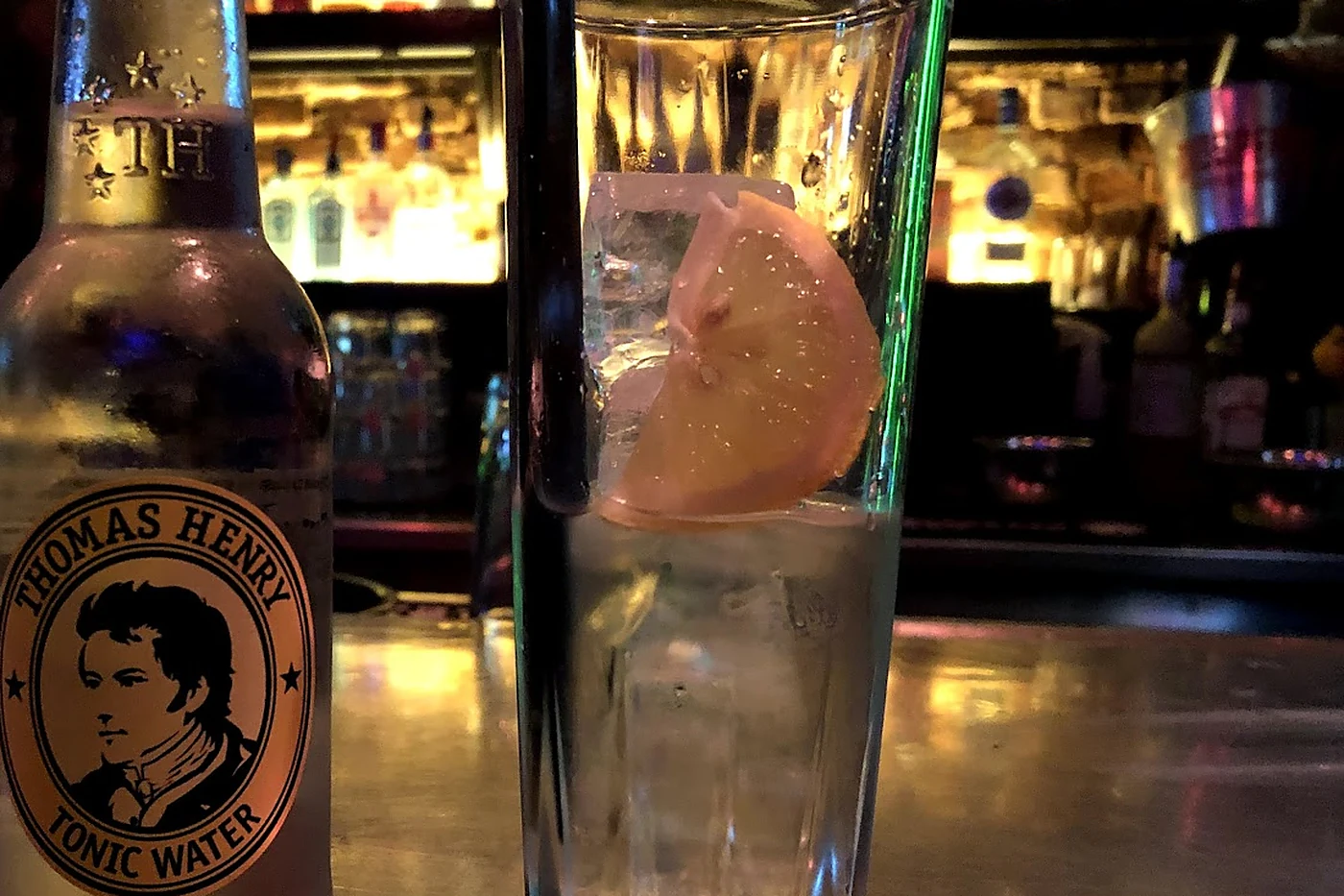 Flasche Thomas Henry Tonic Water neben einem Longdrinkglas mit Eis und Zitrone vor der Flaschenwand