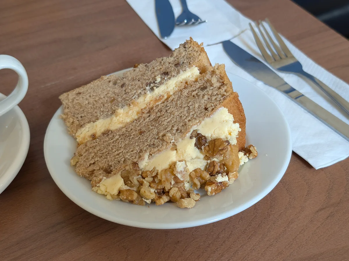 Stück Nusskuchen mit Buttercreme und Walnüssen auf einem weißen Teller, daneben ein Milchkaffee