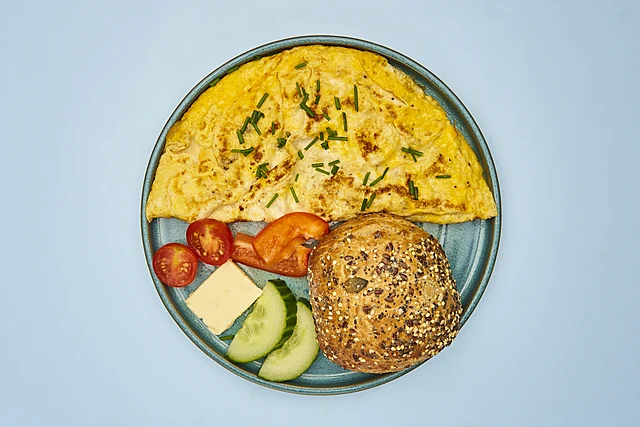 Omelett mit Schnittlauch auf blauem Teller, dazu Körnerbrötchen, Butter, Tomate und Gurke