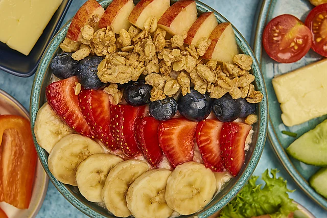 Frühstücksteller von oben mit Banane, Erdbeeren, Blaubeeren, Apfel, Walnüssen und Müsli, daneben Käse und Gurke