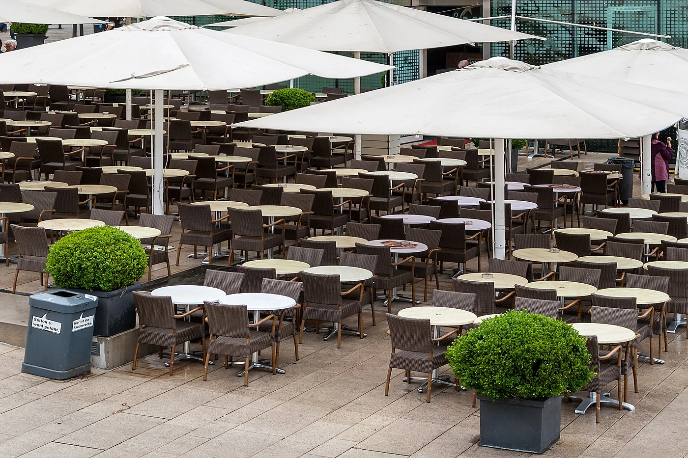 Leere Terrasse des Alsterpavillons mit runden Tischen, Korbstühlen, weißen Sonnenschirmen und Buchsbaumkugeln