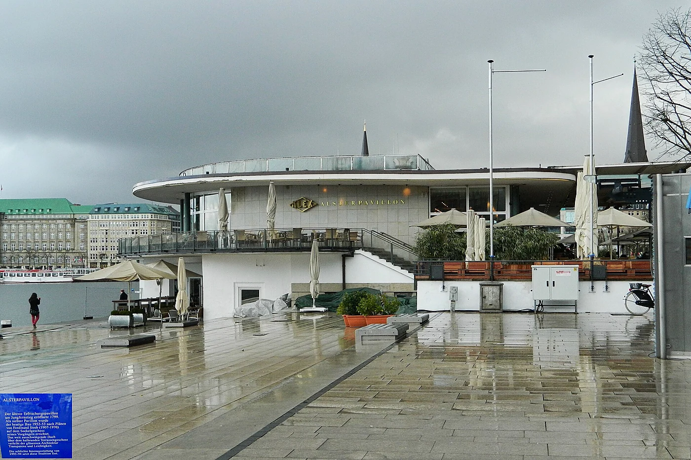 Alsterpavillon von der Seite bei Regen mit nassem Vorplatz, geschlossenen Schirmen und dem Turm der St.-Petri-Kirche im 