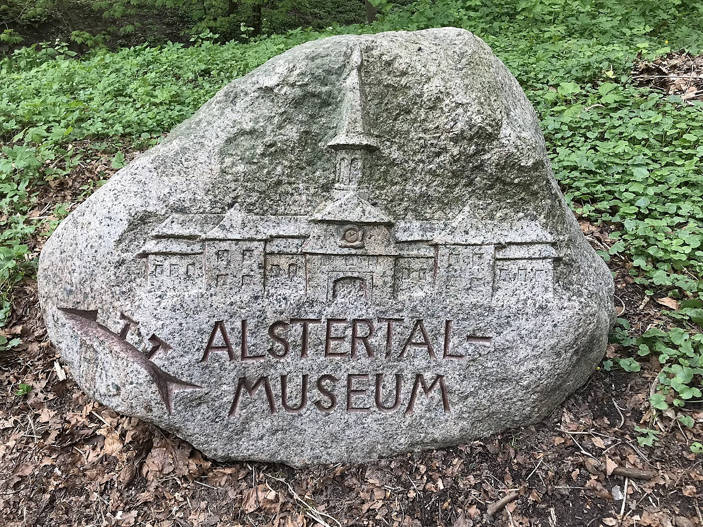 Großer Findling mit eingemeißeltem Torhaus-Relief und der Inschrift „Alstertal-Museum“ zwischen Efeu und Laub