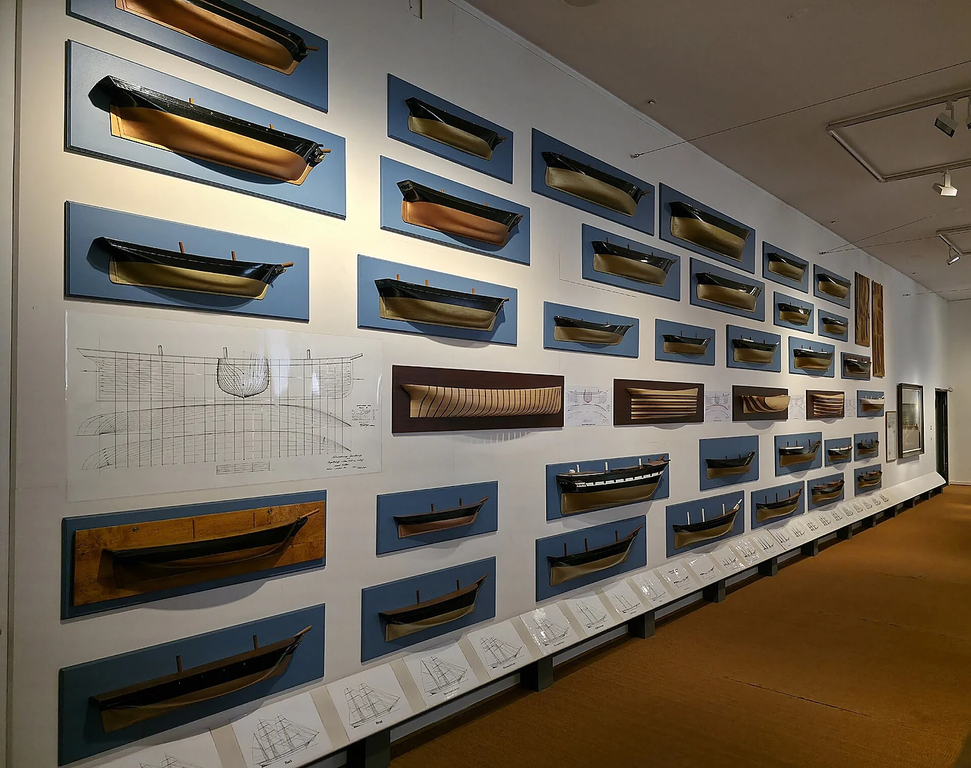 Lange Wand voller Halbmodelle von Schiffsrümpfen auf blauen Tafeln, davor Konstruktionszeichnungen
