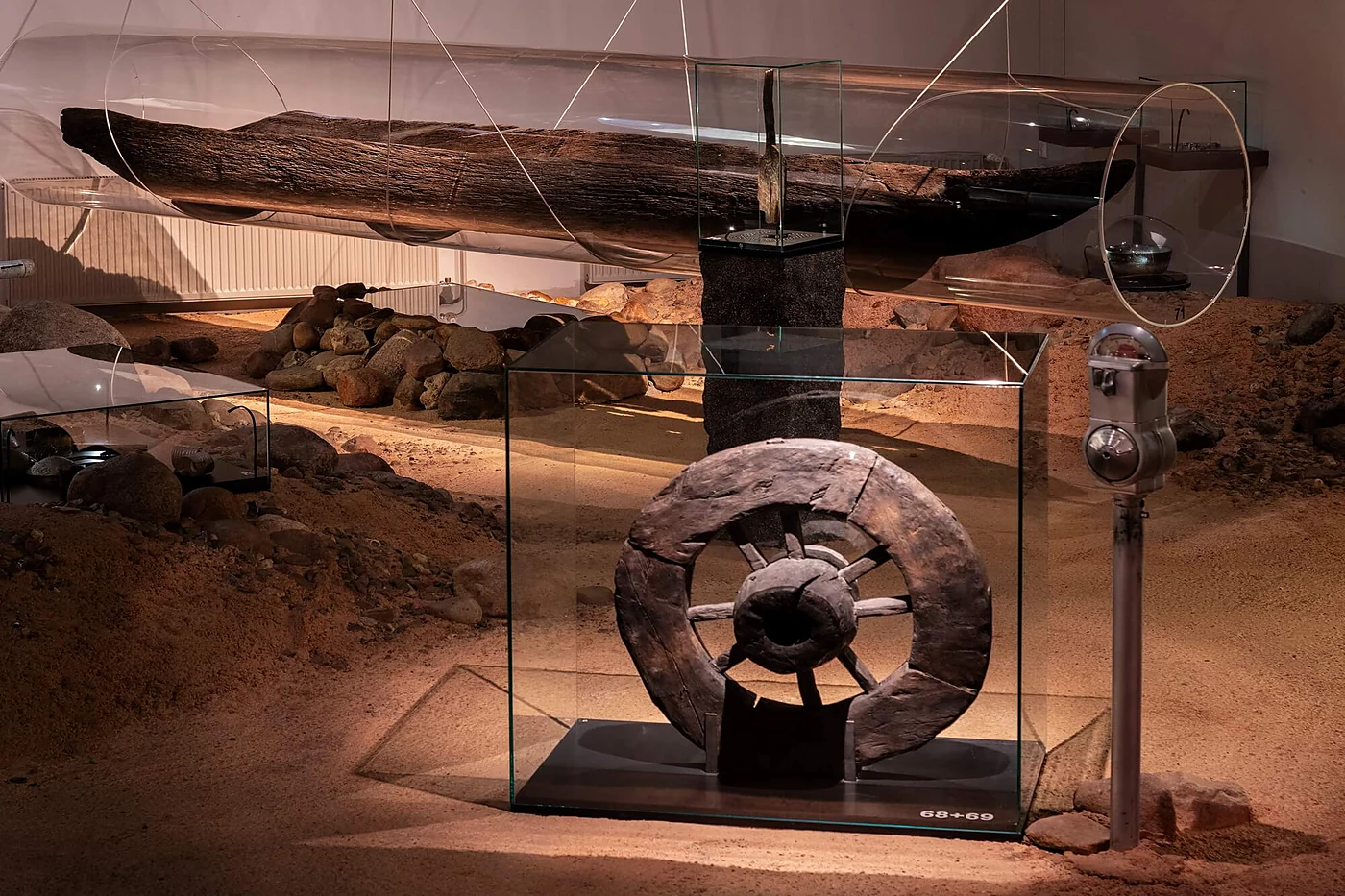 Glasvitrine mit einem alten hölzernen Scheibenrad in einer nachgebauten Ausgrabungslandschaft aus Sand und Findlingen, darüber ein Einbaum unter der Decke