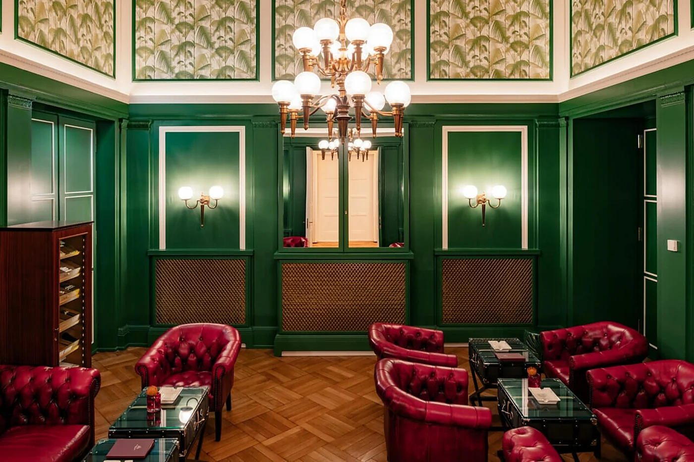 Smoker’s Lounge mit grünen Wandpaneelen, roten Ledersesseln, Kronleuchter und Tapete mit Palmenmuster