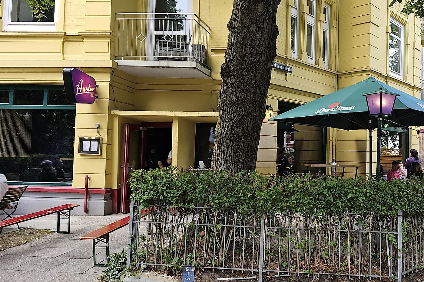 Gelbes Eckhaus mit dem violetten Auster-Bar-Schild, Bierbänken auf dem Bürgersteig und eingezäuntem Biergarten unter einem Baum
