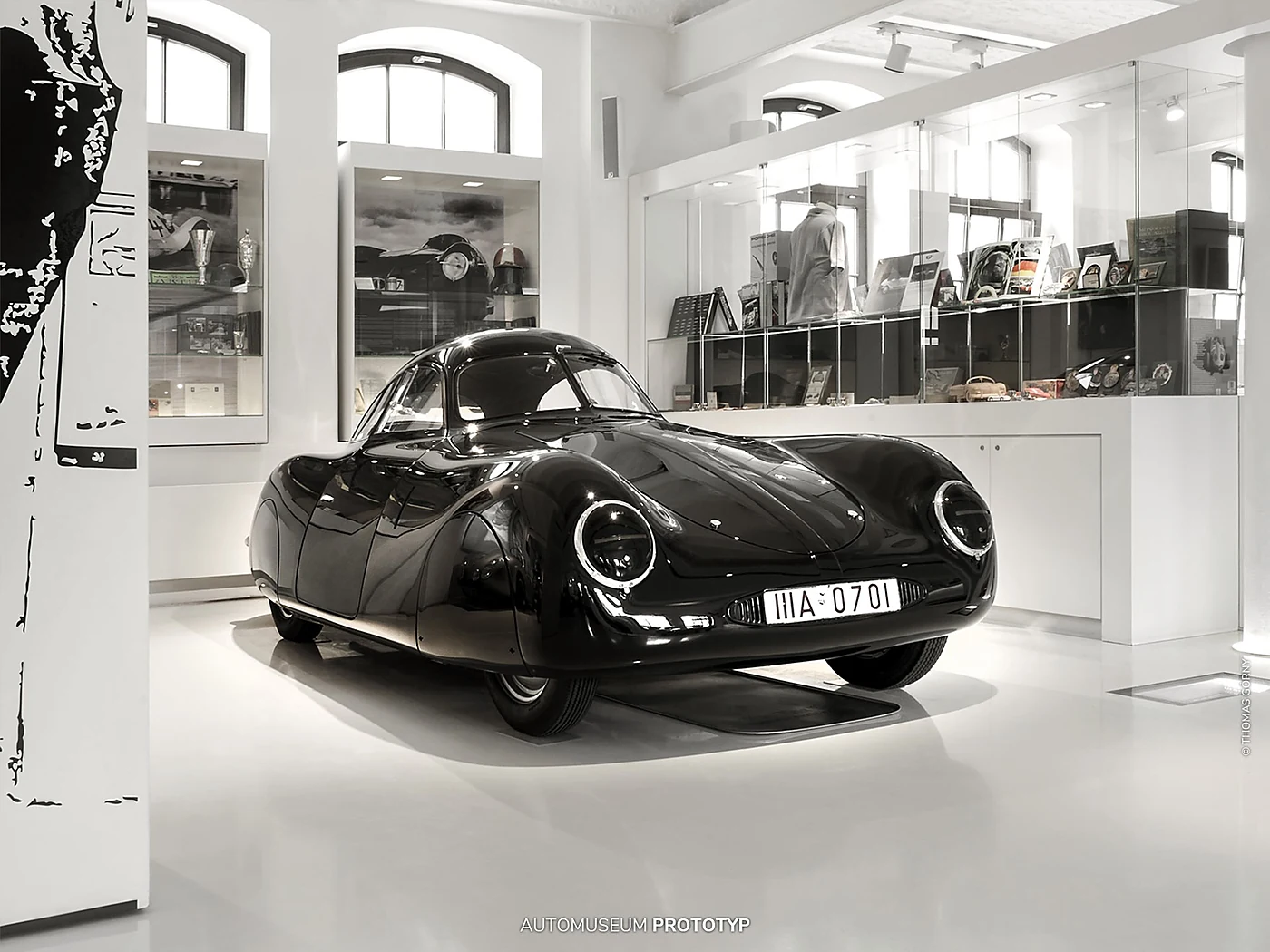 Schwarzer stromlinienförmiger Porsche Typ 64 von 1939 in einem weißen Raum mit Vitrinen und historischen Fotos