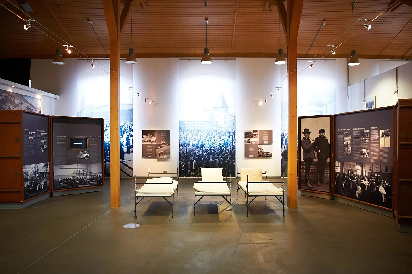 Ausstellungsraum in Haus 1 mit hölzernem Dachstuhl, drei historischen Metallbetten und großformatigen Schwarzweißfotos d