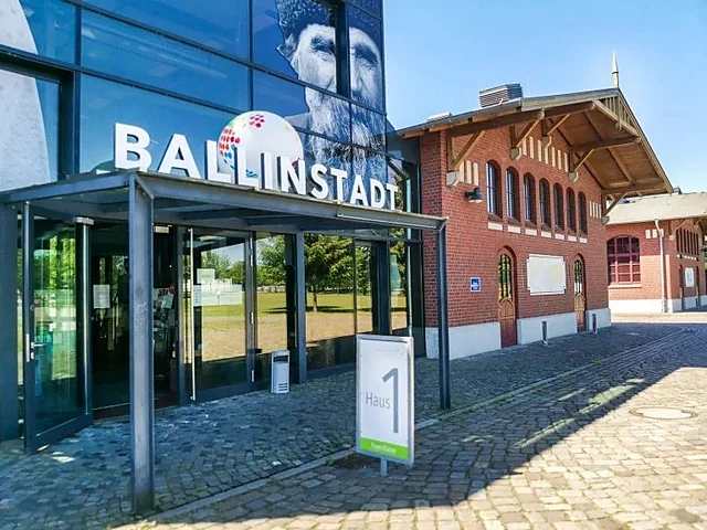 Glaseingang von Haus 1 mit dem Schriftzug BallinStadt, dahinter die roten Backsteinpavillons der nachgebauten Auswandere