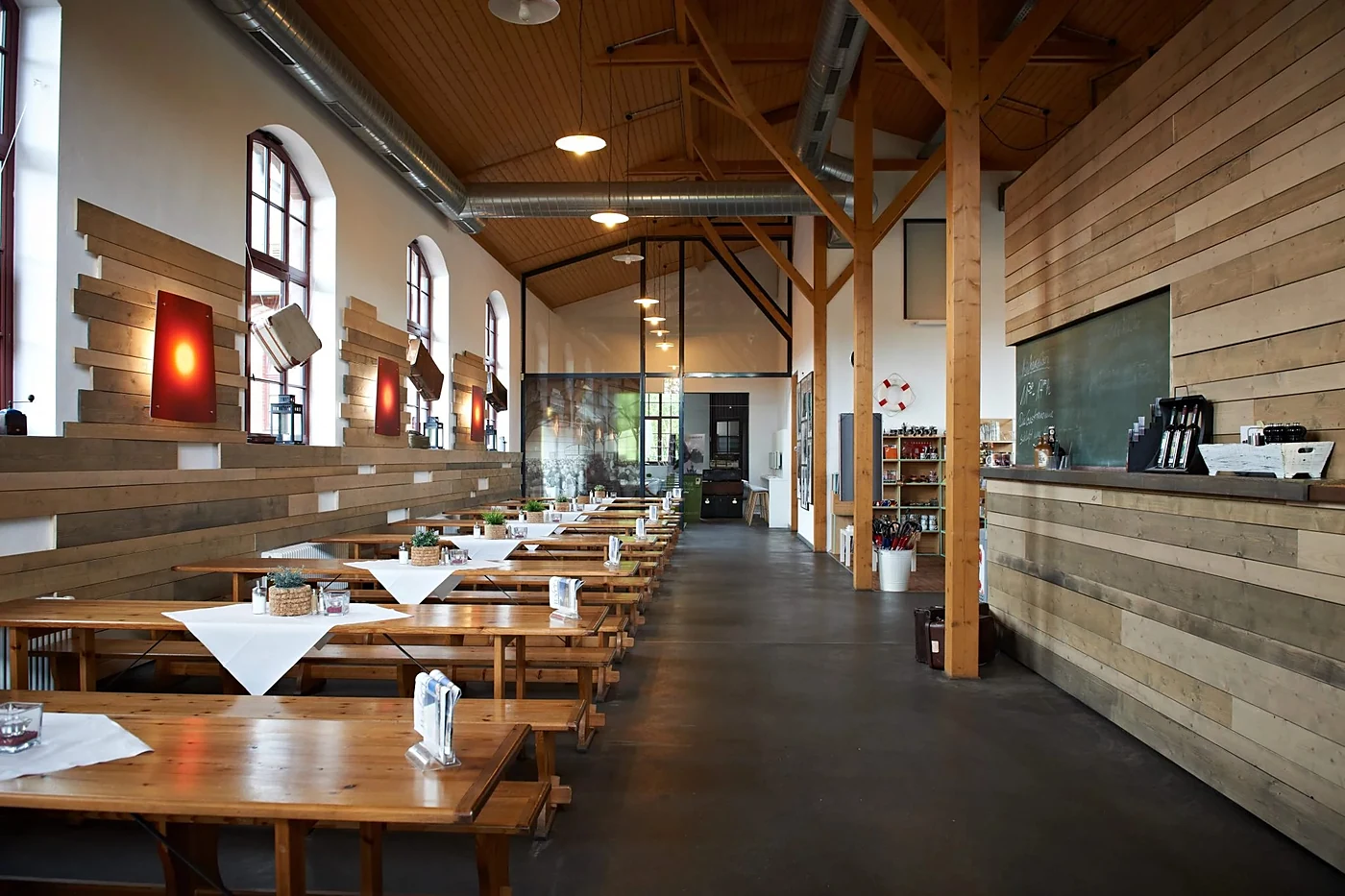 Restaurant Nach Amerika mit langen Holztischen, Bänken, weißen Tischdecken und Holzverkleidung unter dem offenen Dachstu