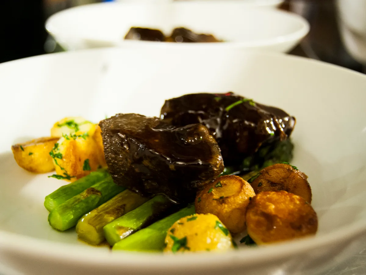 Geschmorte Rinderrippe in dunkler Sauce mit grünem Spargel und Bratkartoffeln in einem weißen Teller