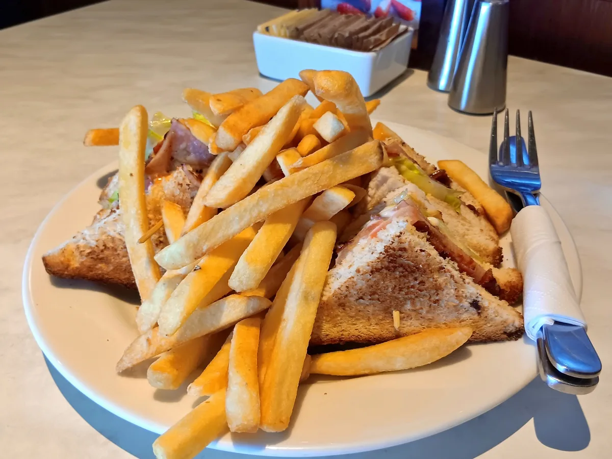 Clubsandwich aus getoastetem Brot mit Fleisch, Salat und Tomate, dazu Pommes frites auf einem hellen Teller