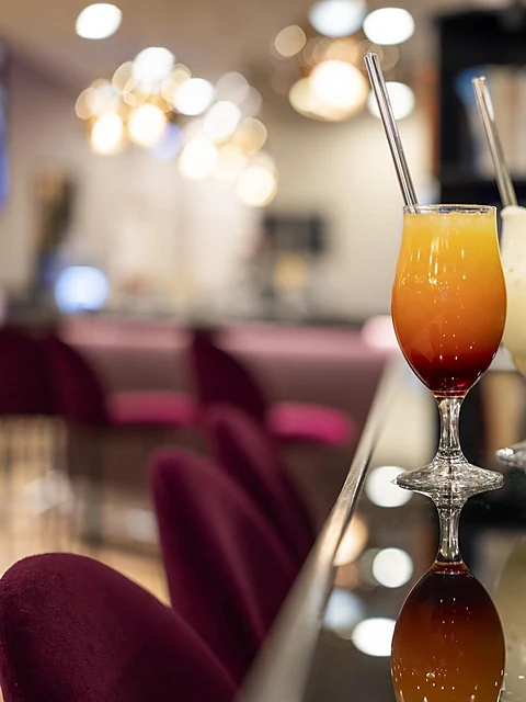 Tequila Sunrise im Stielglas mit Glasstrohhalm auf dem schwarzen Tresen, im Hintergrund weinrote Samthocker und Kugellam
