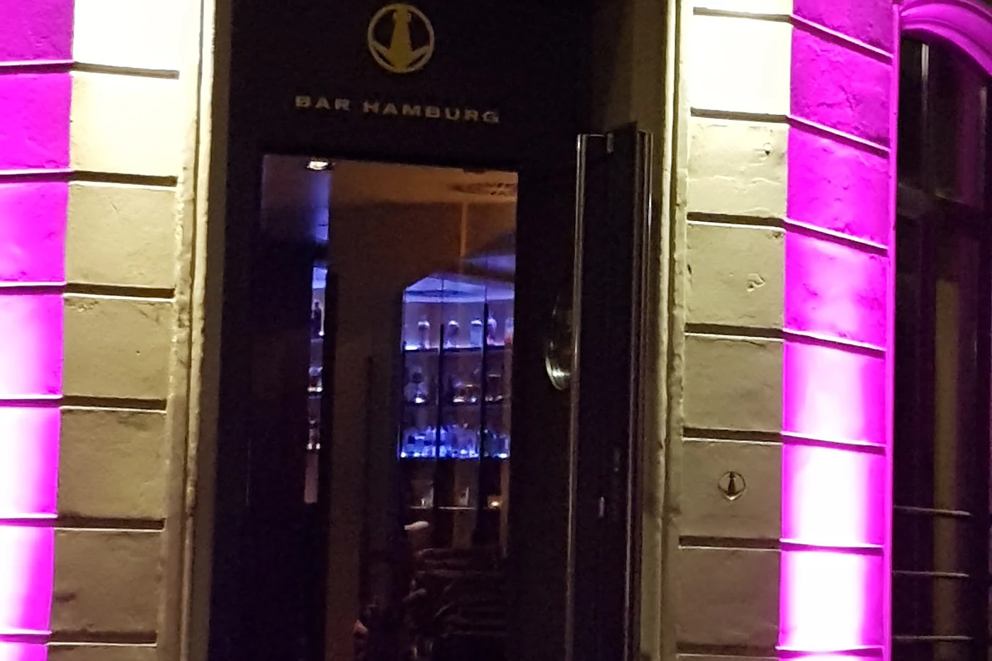 Eingang der Bar Hamburg bei Nacht mit dunkler Tür, Logo, Schriftzug BAR HAMBURG und violett angestrahlter Fassade