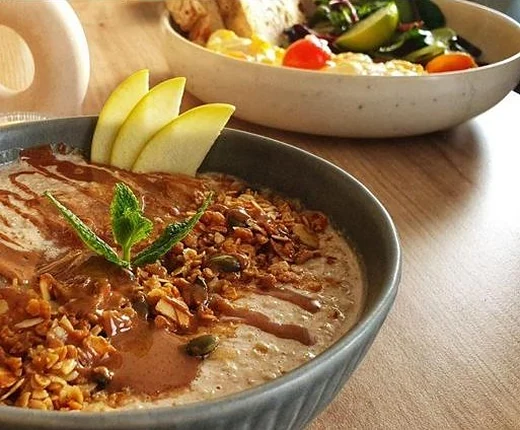 Overnight Oats mit Mandelmus, Granola, Apfelspalten und Minze in einer grauen Schale