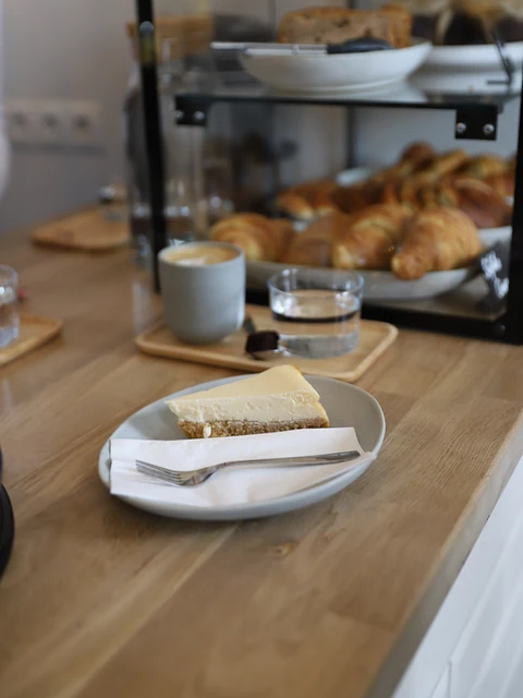 Stück Karottenkuchen auf einem Teller, dahinter Cappuccino und Croissants in der Vitrine