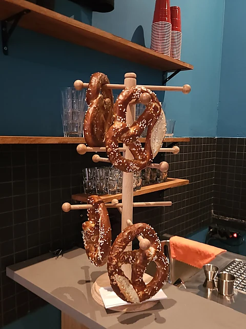 Laugenbrezeln hängen an einem Holzständer auf dem Tresen vor türkisfarbener Wand und Regalen