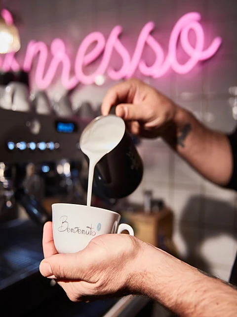 Barista gießt aufgeschäumte Milch in eine Benvenuto-Tasse, im Hintergrund die pinke Neonschrift Espresso