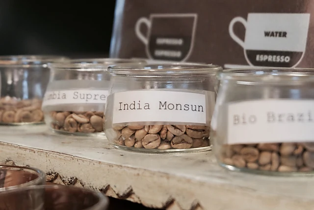 Gläser mit rohen und gerösteten Kaffeebohnen, beschriftet mit Sorten wie India Monsun und Bio Brazil