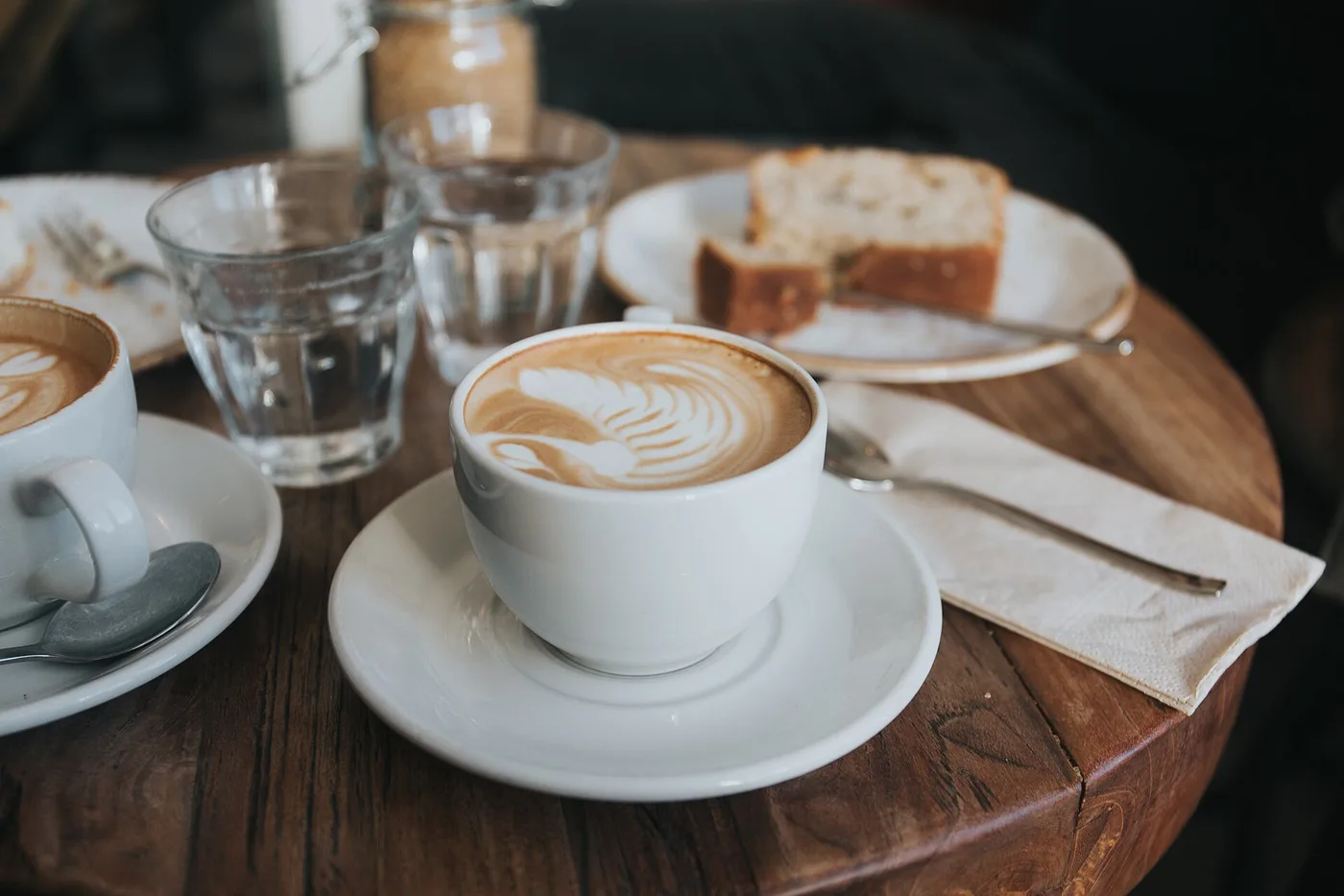 Cappuccino mit Milchschaummuster, daneben ein Stück Kuchen und Wassergläser auf einem Holztisch