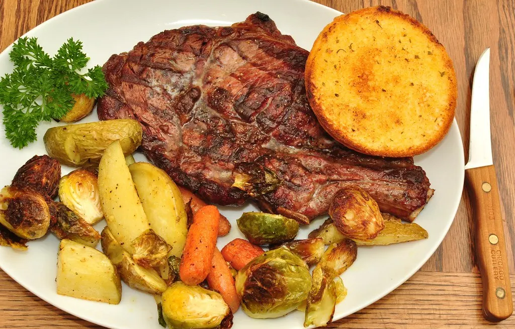 Gegrilltes Steak mit Röstkartoffeln, Rosenkohl, Möhren und geröstetem Brot auf weißem Teller