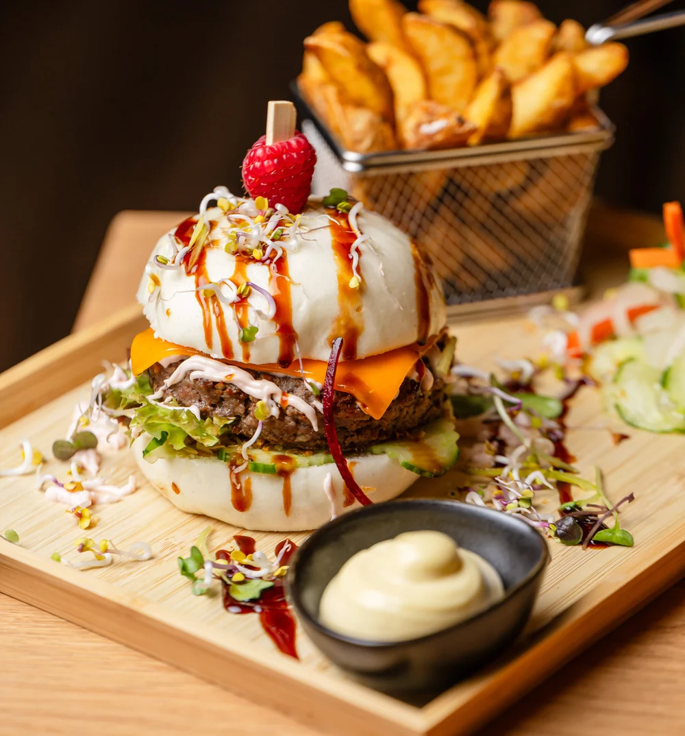 Bao Burger mit Rinderpatty, Käse, Salat und Sprossen im gedämpften Bun, dahinter Kartoffelwedges im Drahtkorb und Dip