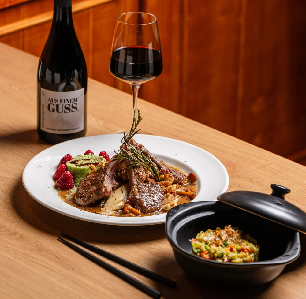 Gegrilltes Lammkotelett mit Rosmarin und Himbeeren auf weißem Teller, dazu gebratener Reis im Tontopf und Rotwein