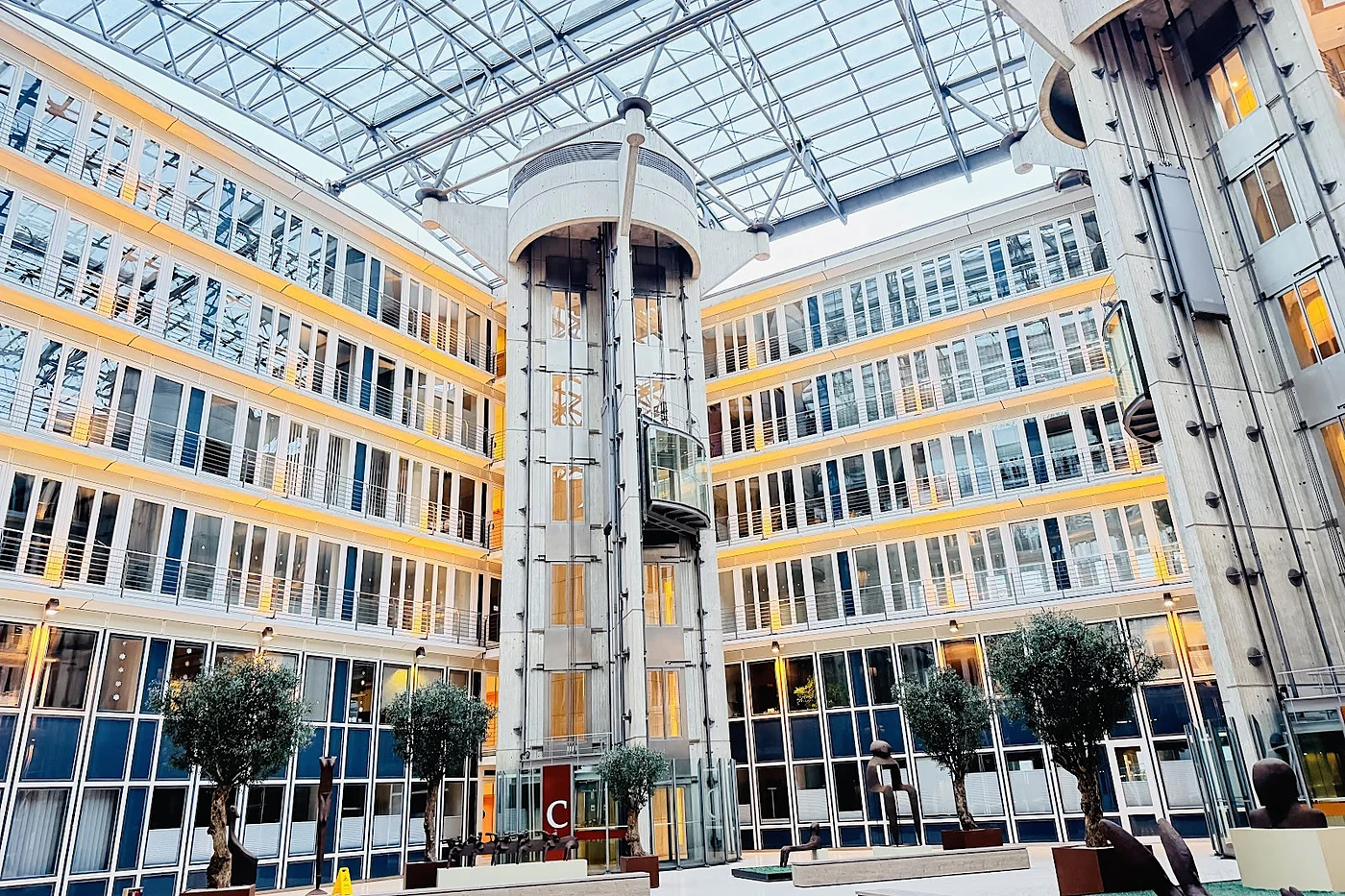 Verglastes Atrium eines Bürogebäudes mit Stahl-Glas-Dach, Aufzugstürmen und Olivenbäumen im Innenhof
