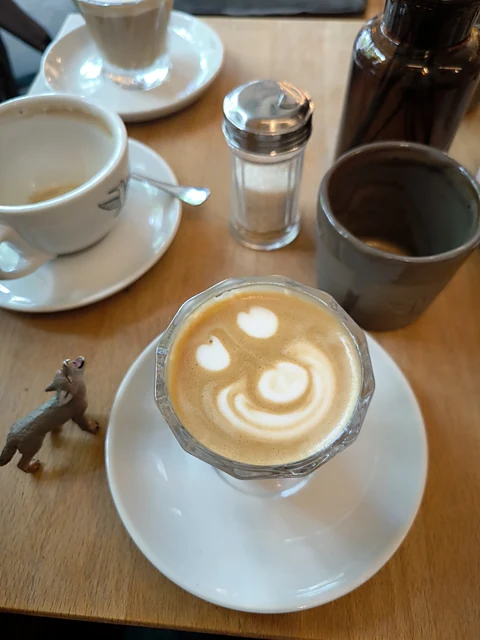 Cappuccino mit Latte-Art im Glas auf weißer Untertasse, dahinter eine Kaffeetasse, Zuckerstreuer und ein Espresso-Becher auf Holztisch