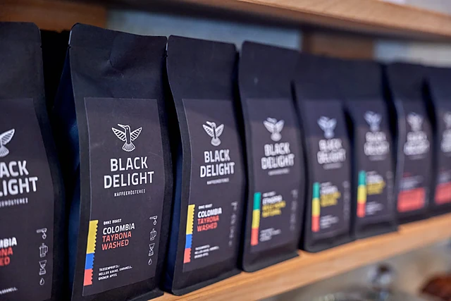 Reihe schwarzer Kaffeetüten mit Kolibri-Logo und bunten Etiketten, etwa Colombia Tayrona Washed, auf einem Holzregal