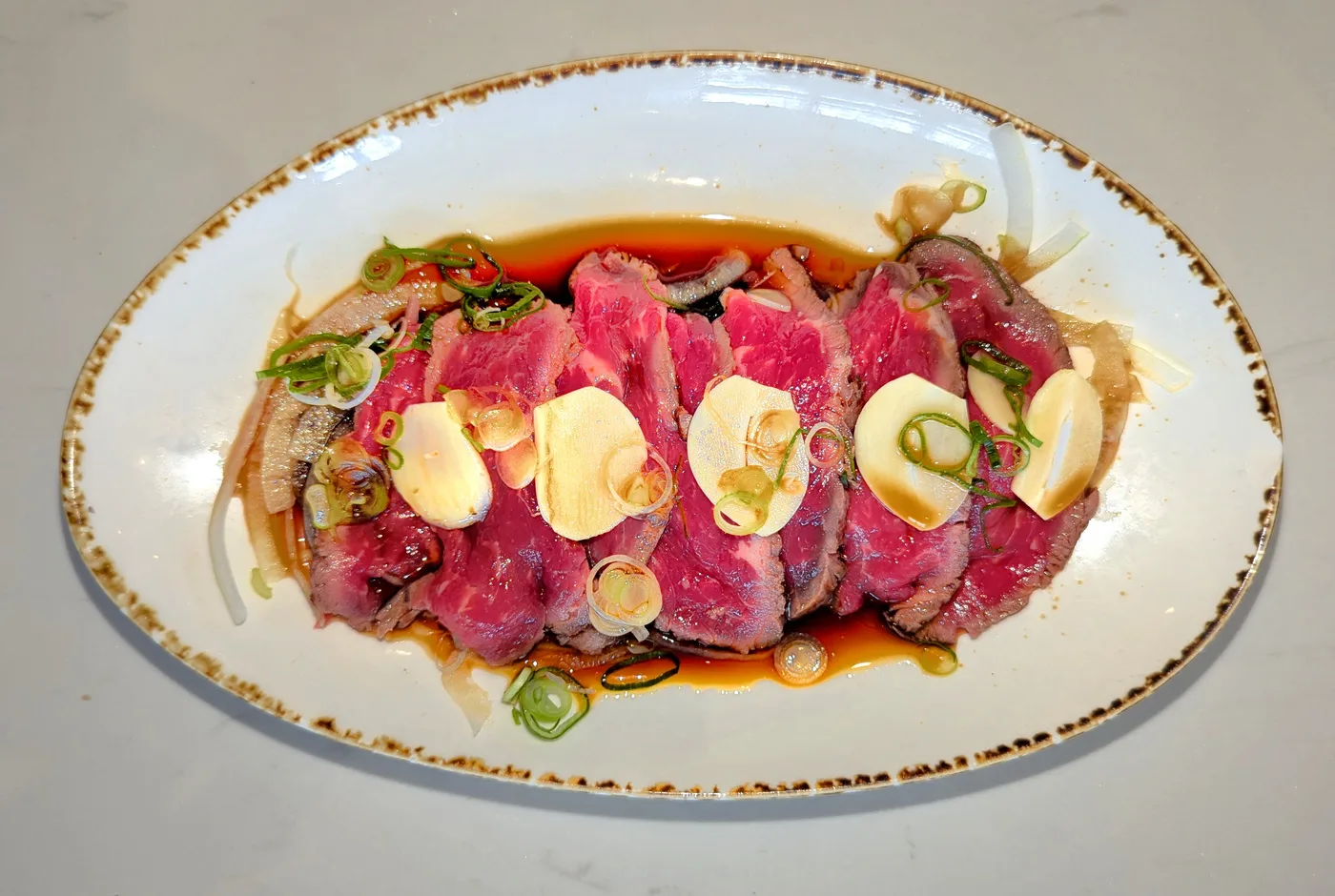 Dünn geschnittenes Rinderfilet in Ponzu mit Frühlingszwiebeln und Knoblauchchips auf einem ovalen Teller