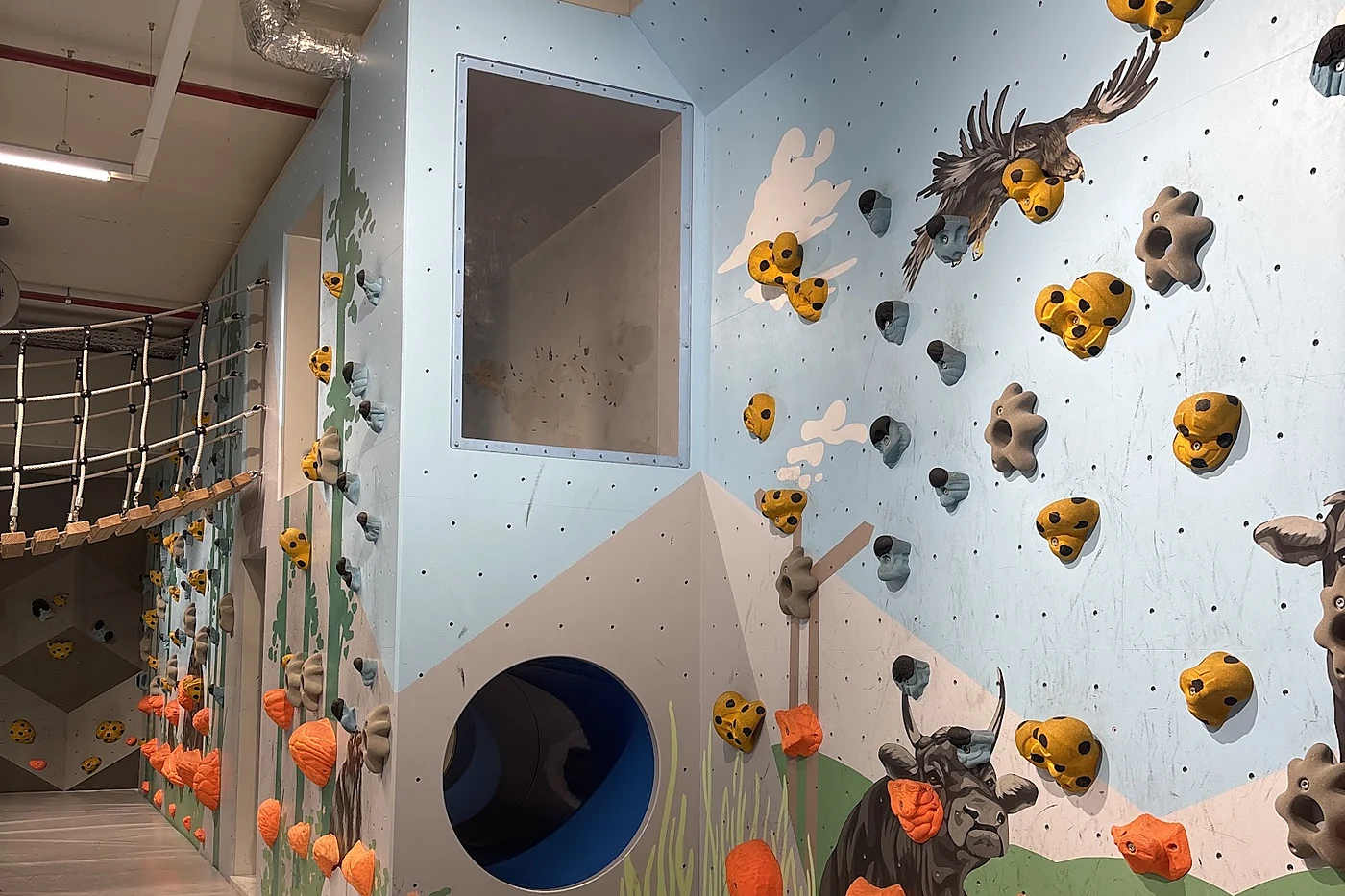 Kinderbereich mit hellblauer Kletterwand, aufgemalten Tieren, gelben Griffen und einem runden Tunnel in der Wand