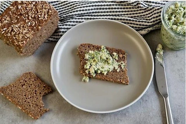 Scheiben dunkles Körnerbrot, eine davon mit Kräuterfrischkäse auf einem grauen Teller