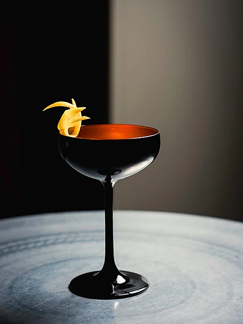 Dunkler Cocktail in einer schwarzen Coupette mit Orangenzeste auf einem Marmortisch