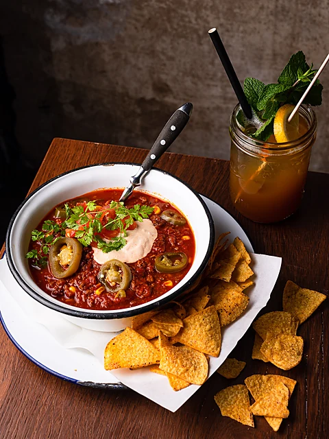 Chili in einer weißen Schale mit Nachos, daneben ein Cocktail im Glas