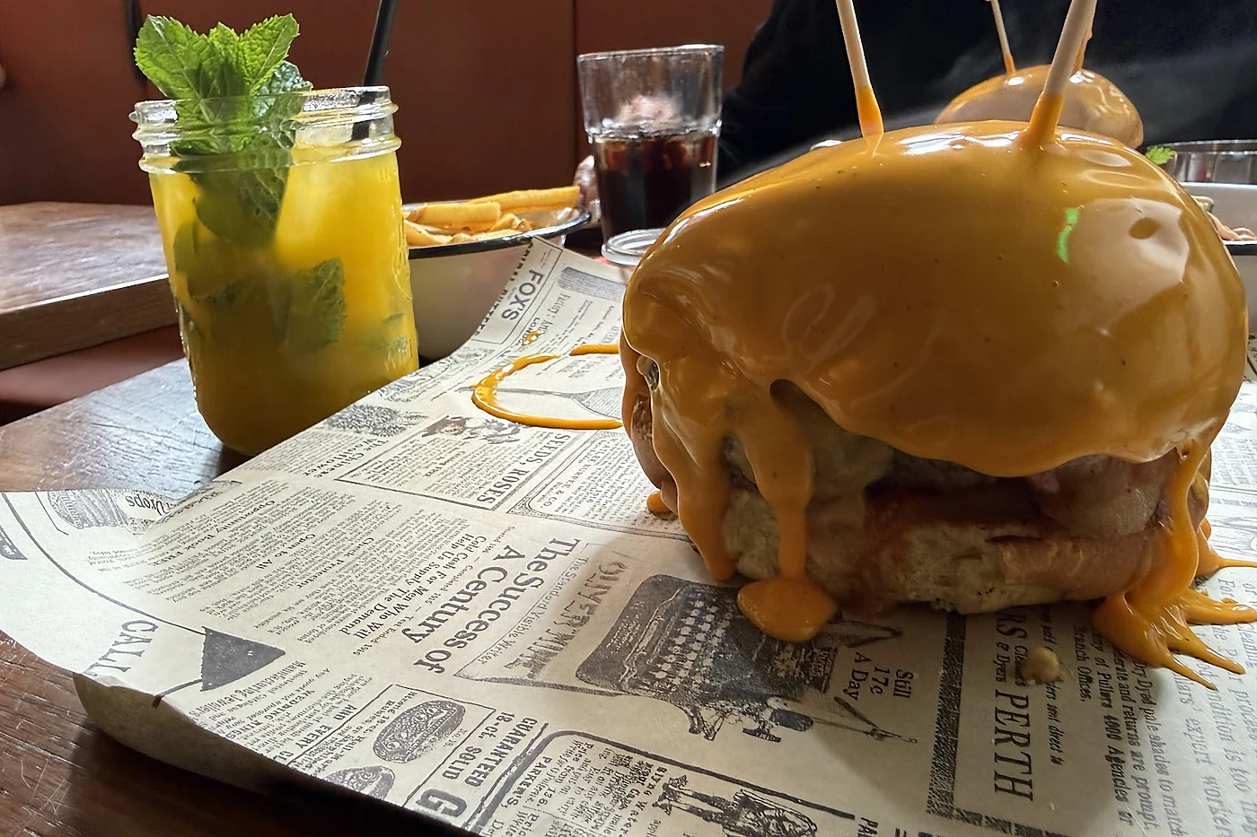 Burger mit übergossener Käsesauce auf Zeitungspapier, daneben ein Cocktail