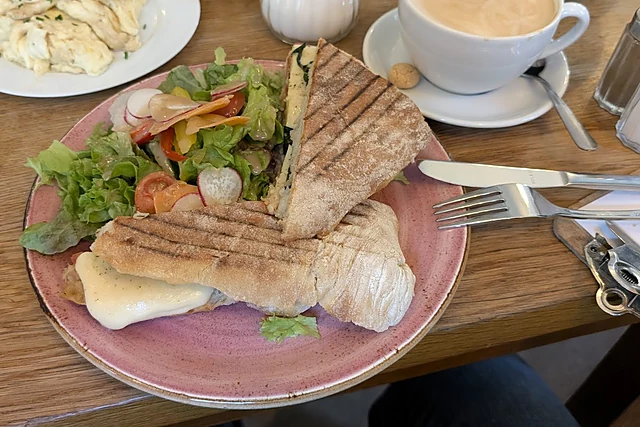 Gegrilltes Panini mit Salat und Tomaten auf rosa Teller, dahinter Rührei, Brötchen und ein Milchkaffee auf Holztisch