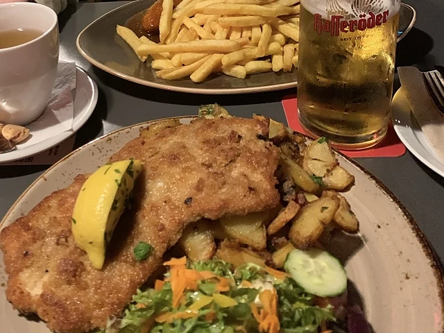 Paniertes Schnitzel mit Zitrone, Bratkartoffeln und Salat auf braunem Teller, dahinter Pommes frites und ein Glas Bier