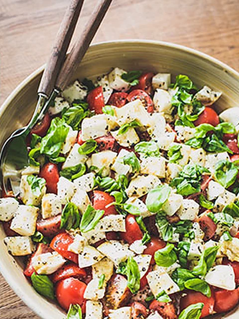 Salatschüssel mit Tomaten, Mozzarella, Basilikum und Brotwürfeln, dahinter Salatbesteck