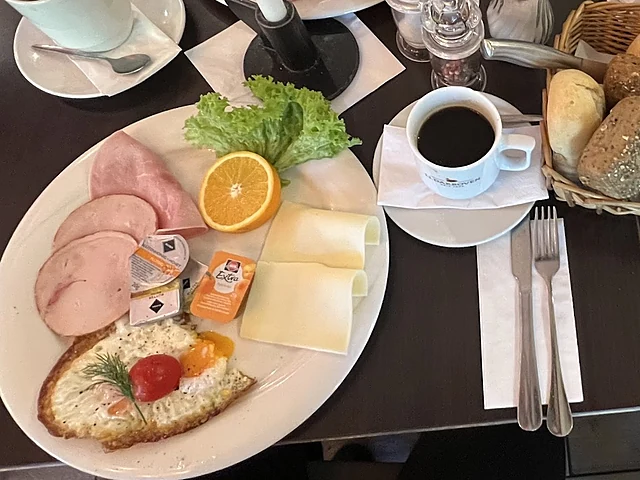 Frühstücksteller mit Schinken, Käse und Orangenscheibe, daneben Kaffee und ein Brötchenkorb auf dunklem Tisch