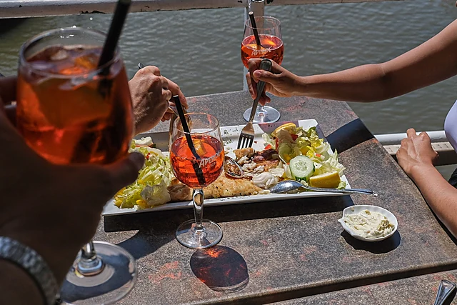 Zwei Hände stoßen mit Aperol Spritz an, darunter eine Fischplatte mit Salat, Zitrone und Remoulade an der Elbe