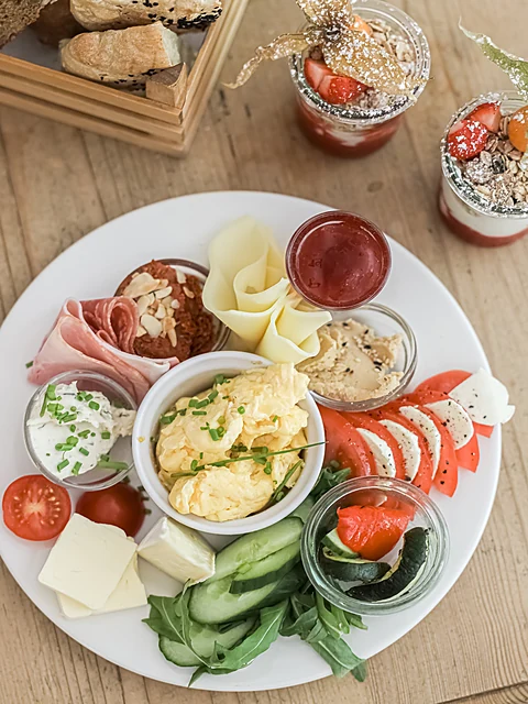 Frühstücksteller von oben mit Rührei, Schinken, Käse, Tomate-Mozzarella, Gurke, Aufstrichen und Marmelade, dahinter Brot