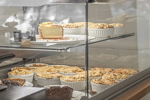 Kuchenvitrine mit vielen kleinen Crumbles in weißen Förmchen und einem angeschnittenen Kuchen