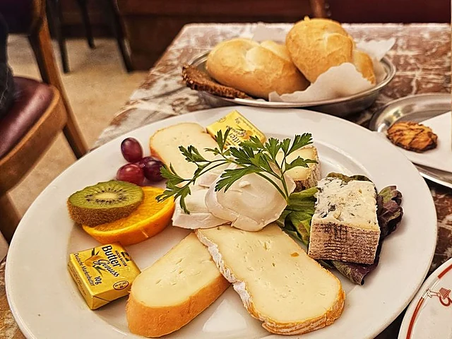 Frühstücksteller mit Käseauswahl, Kiwi, Orange und Trauben, dahinter Brötchenkorb und ein Milchkaffee auf Marmortisch