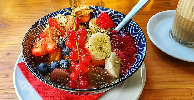 Schale mit Porridge, Erdbeeren, Blaubeeren, Banane, Johannisbeeren und Physalis, daneben ein Latte