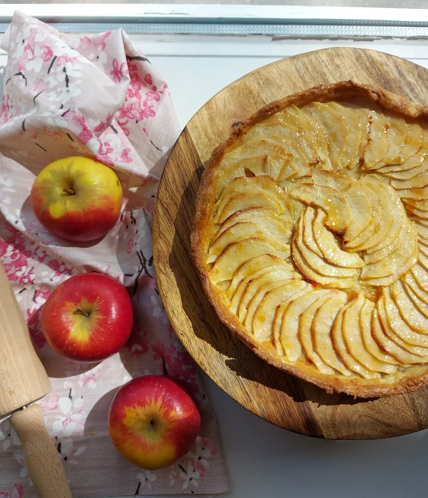 Apfeltarte mit fächerförmig gelegten Apfelscheiben auf einem Holzbrett, daneben frische Äpfel