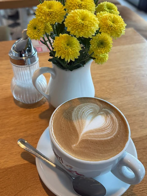 Cappuccino mit Milchschaumherz auf einem Holztisch, daneben gelbe Blumen in einer Vase